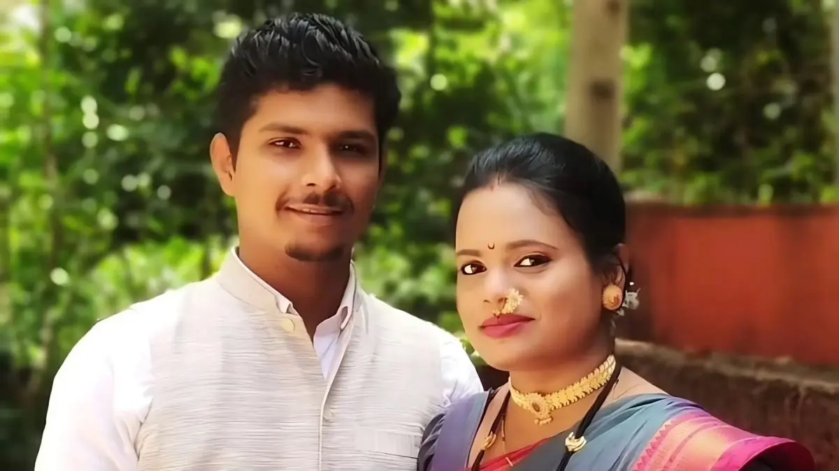 A couple in colorful traditional clothes stand together outside in a green Indian village setting.
